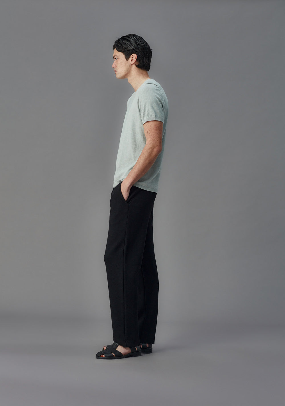 Man wearing a light blue t-shirt and black pants standing against a gray background