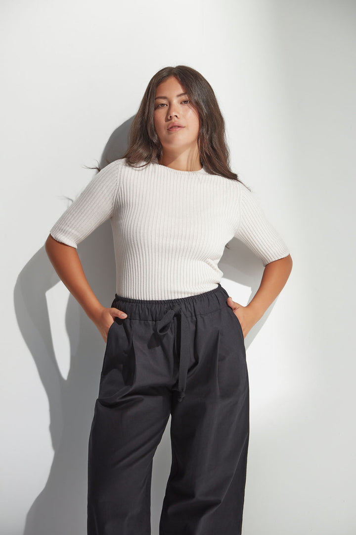 Woman wearing a white ribbed sweater and dark pants against a white background