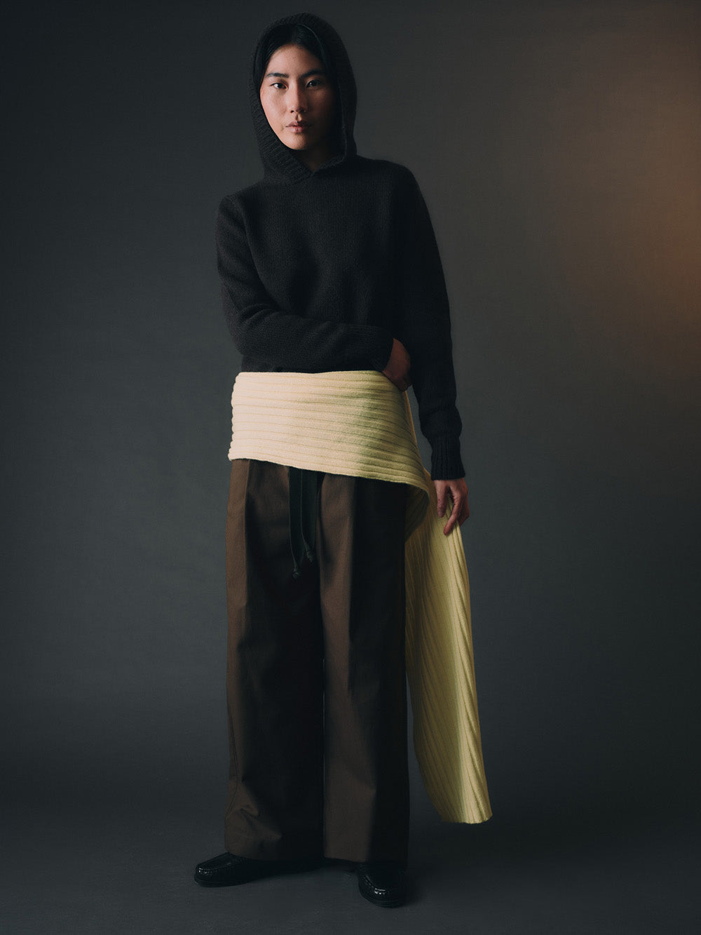 Person wearing a black hooded knit sweatshirt and yellow scarf against a dark background