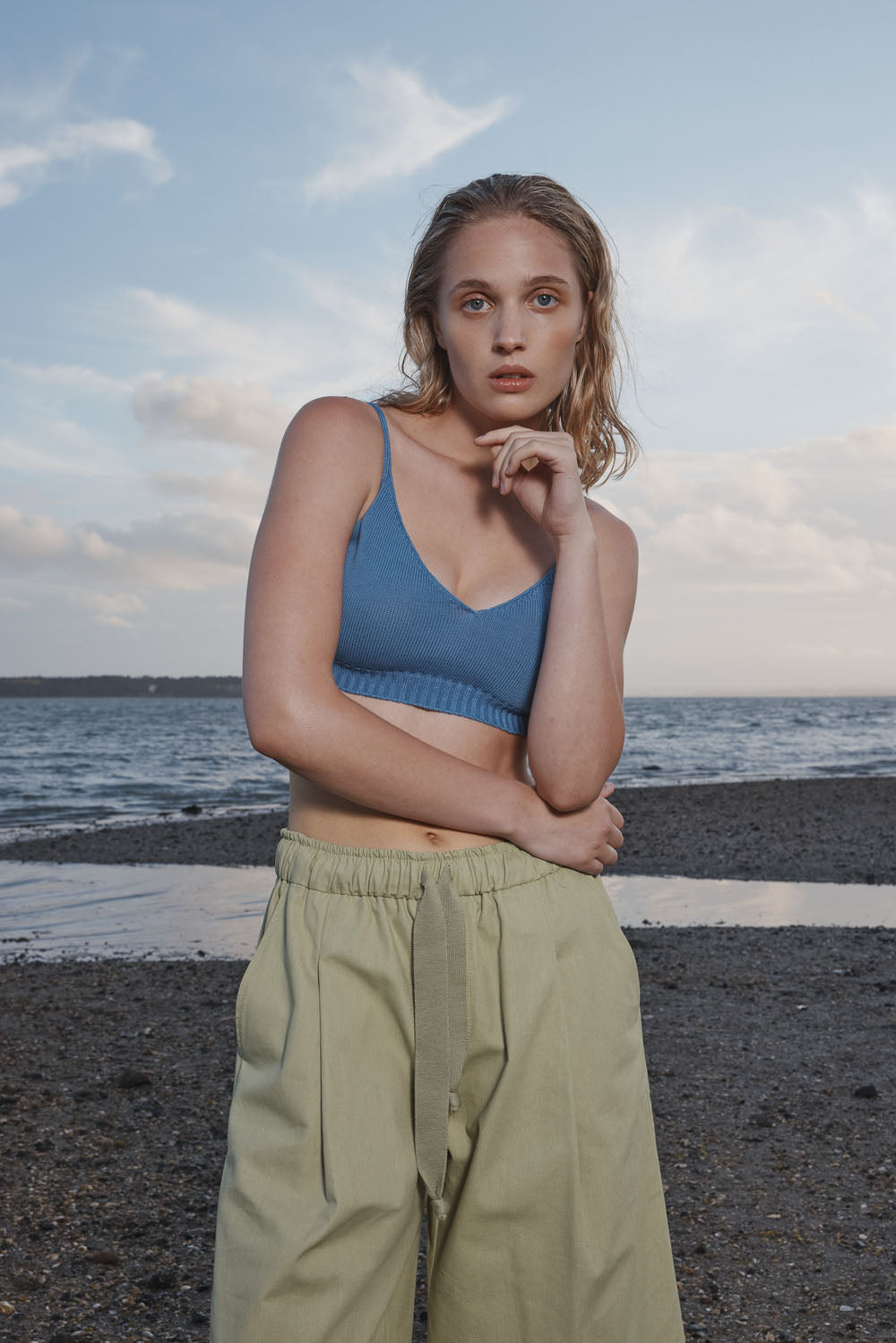 Woman standing on a beach wearing a blue knit bralette and light green pants.