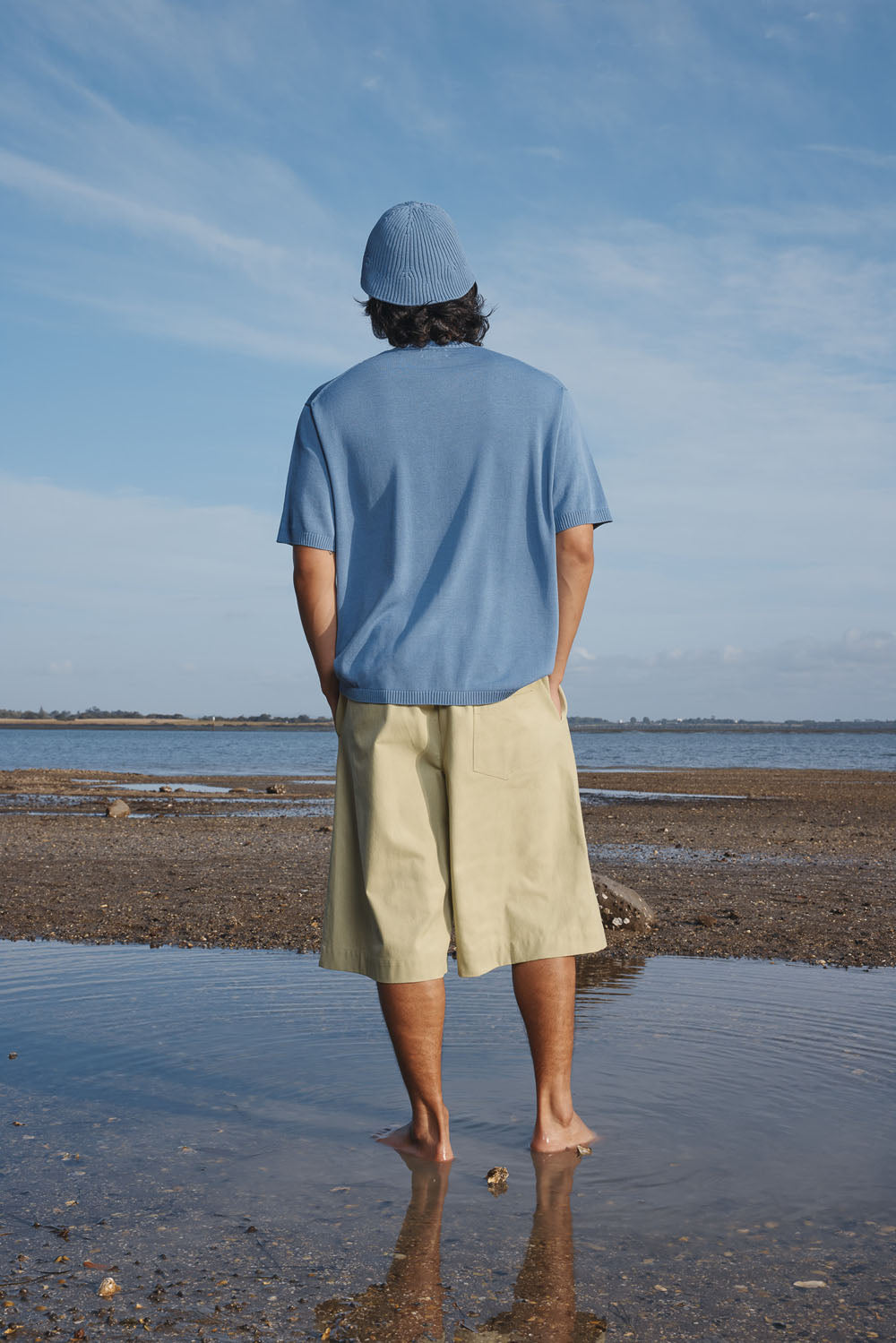 Man wearing a blue shirt and hat and light green shorts standing on a beach with a clear sky.