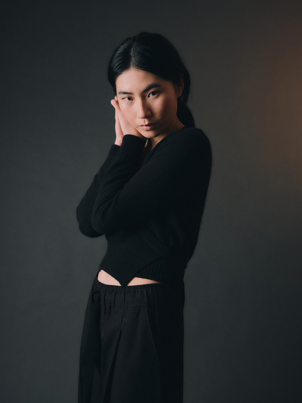 Woman wearing a black knitted wrap sweater and black wide-leg pants against a dark background