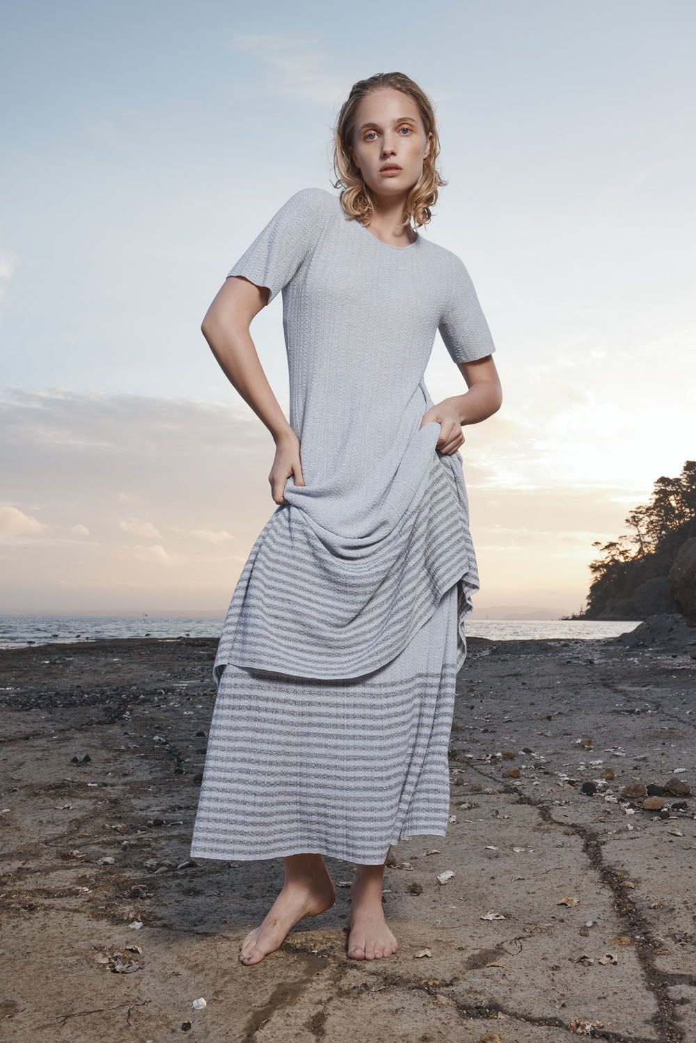Woman in a light gray dress standing on a beach at sunset.