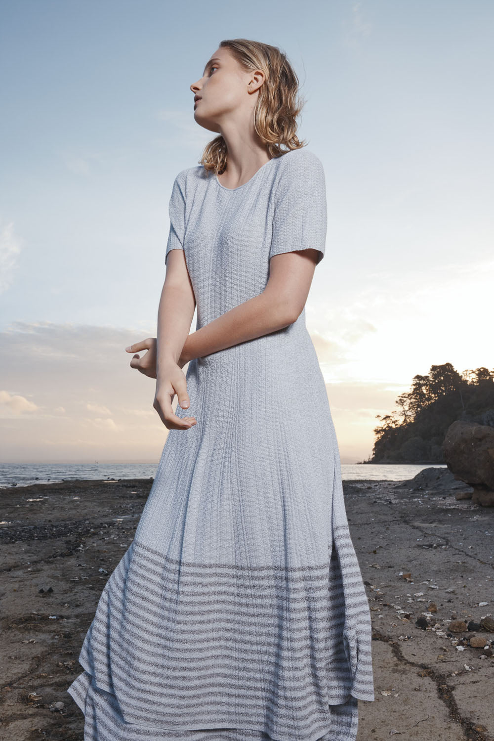 Woman in a light grey dress standing on a beach with a sunset in the background.