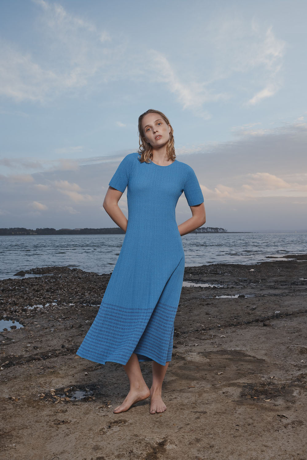 Woman in a blue dress standing on a rocky beach with ocean and sky in the background.