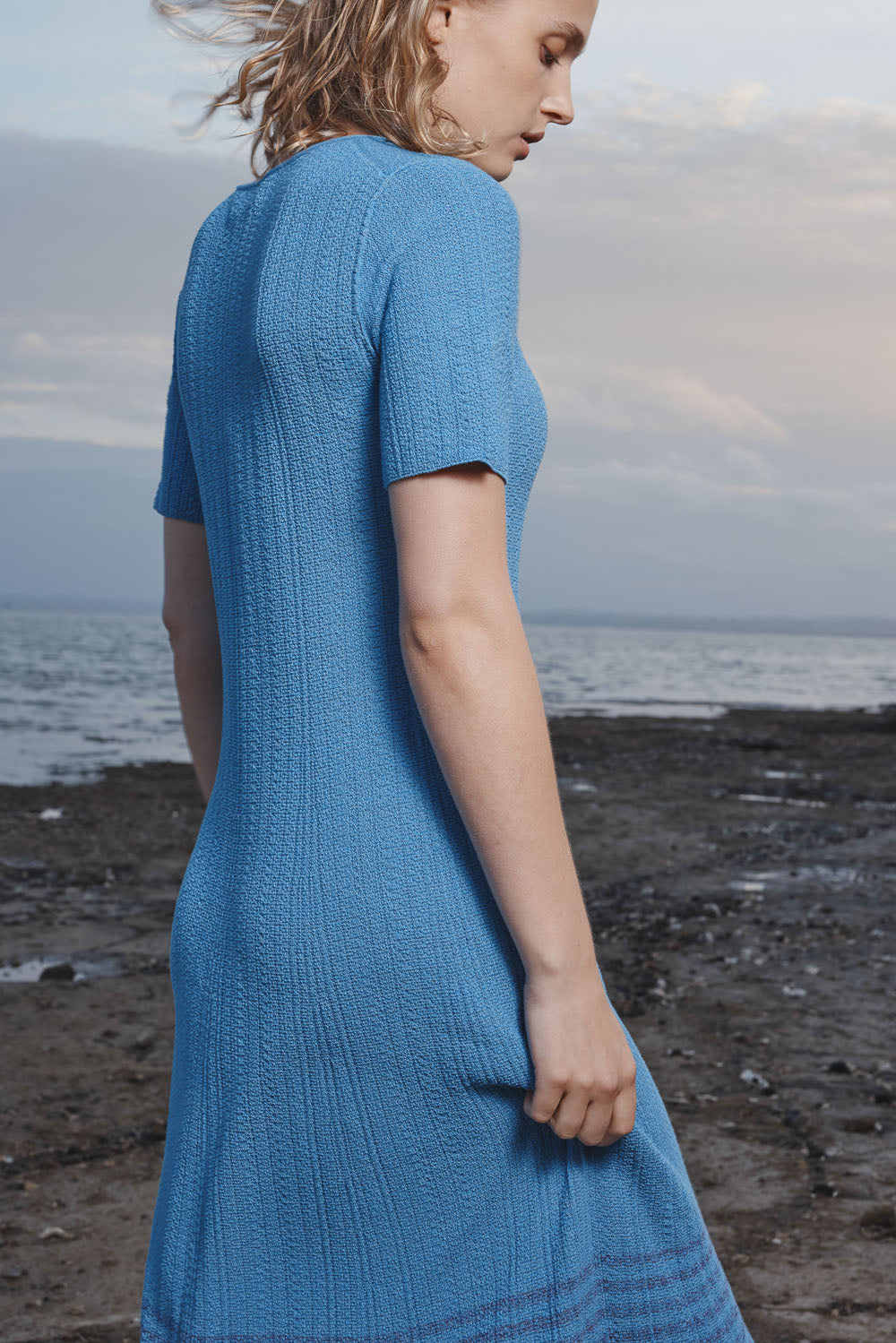 Woman wearing a blue dress standing on a beach with ocean and sky in the background.