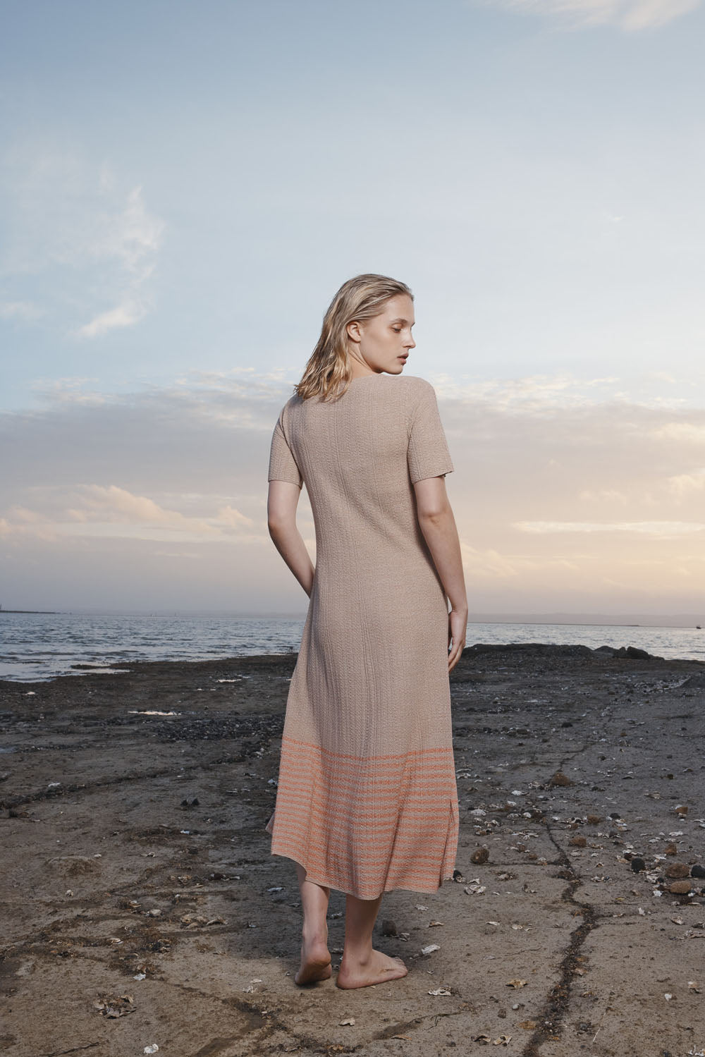 Woman in a beige dress with her back to the camera standing on a beach with a cloudy sky.