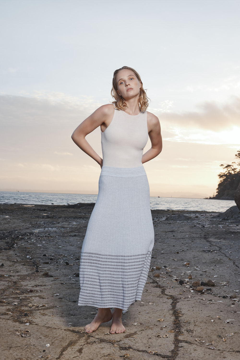 Woman in a grey skirt and white top standing on a beach at sunset.