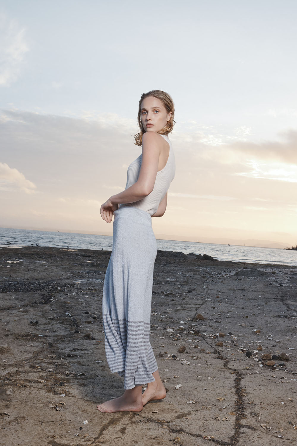 Woman in a grey skirt and white top standing on a beach at sunset.