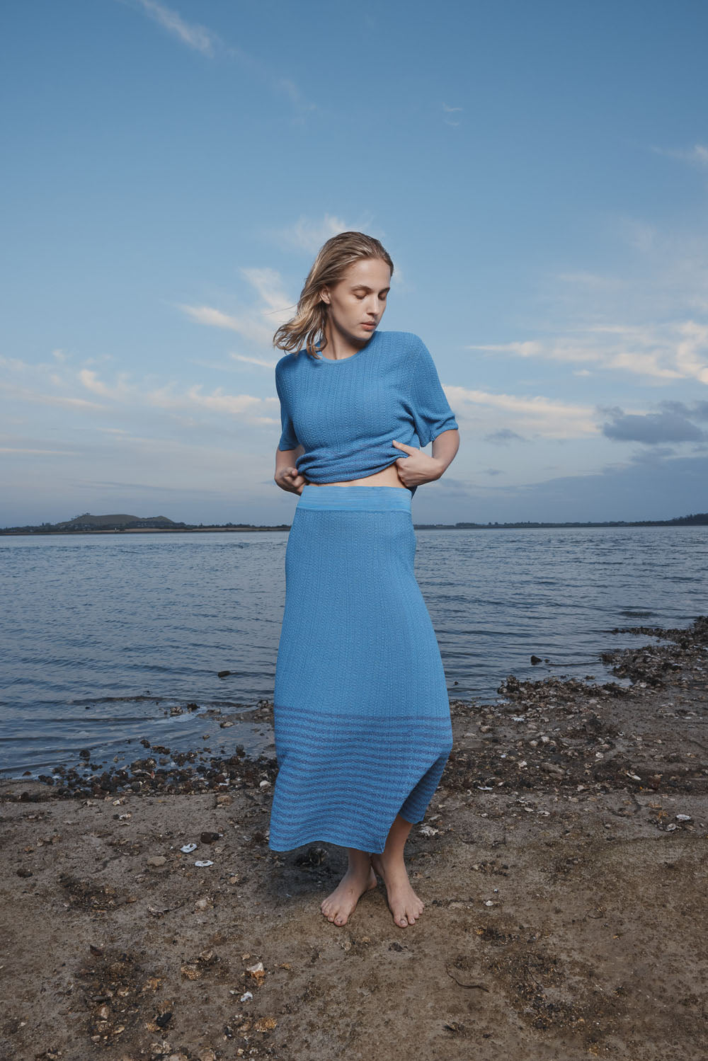 Woman in a blue skirt and top standing on a beach with a body of water and sky in the background.