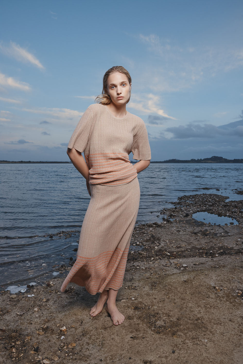 Woman in a beige skirt and top standing by a body of water with a blue sky.