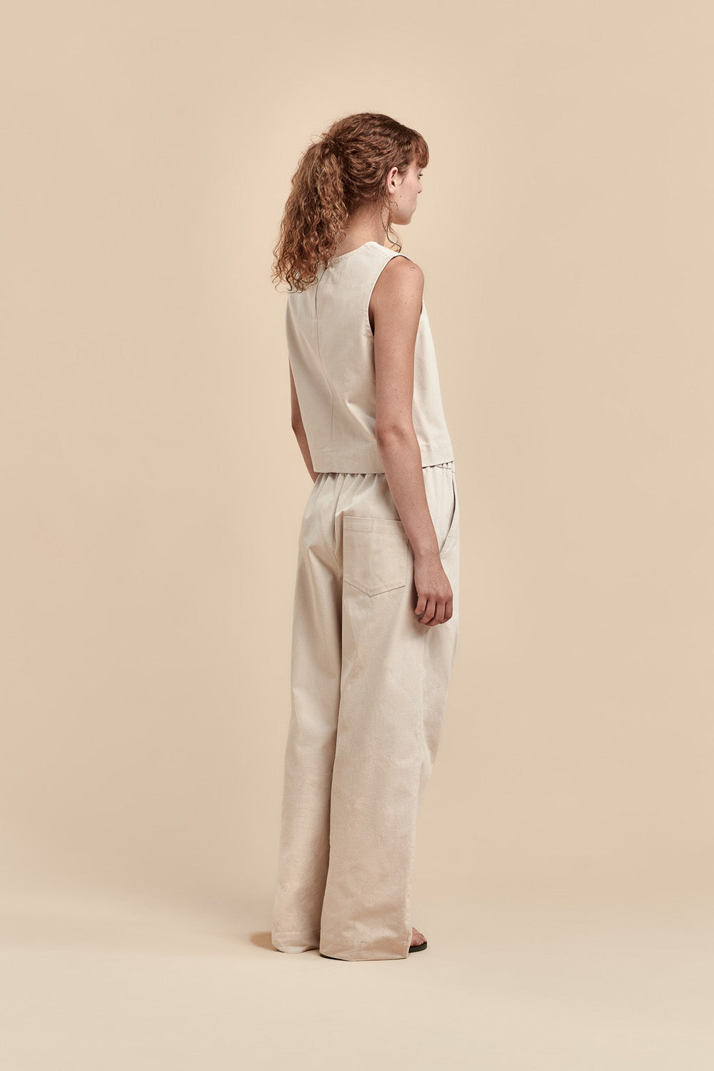 A woman stands in profile wearing a sleeveless cream top and matching wide-leg trousers against a plain beige studio background.