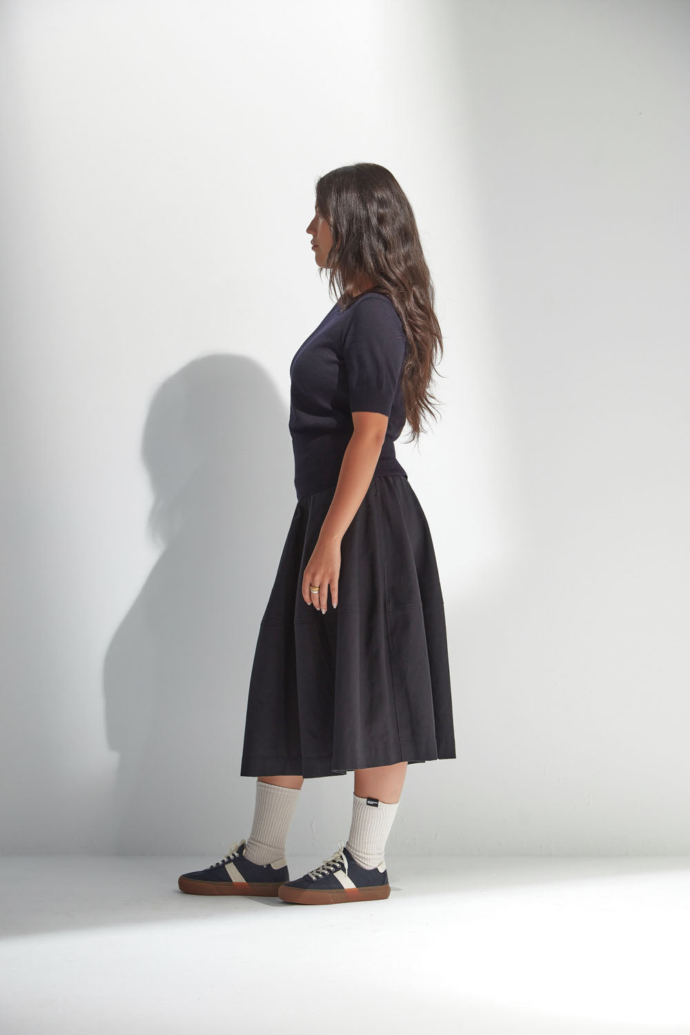 Woman wearing a black skirt and top standing against a white wall.