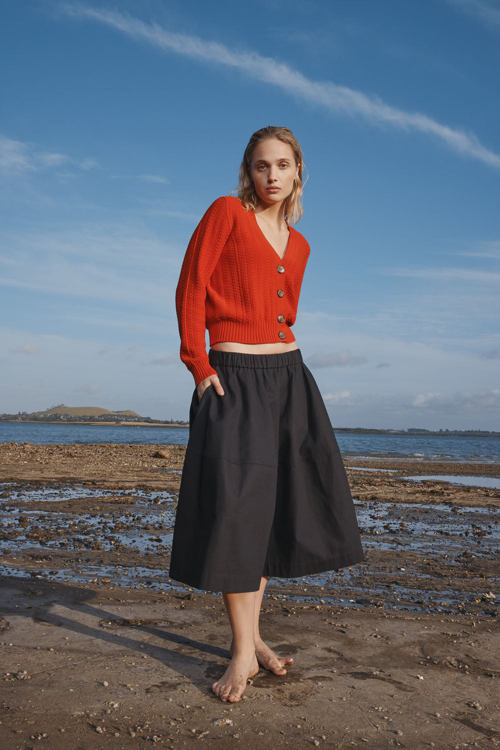 Woman in a red cardigan and black skirt standing on a beach with blue sky and water in the background.