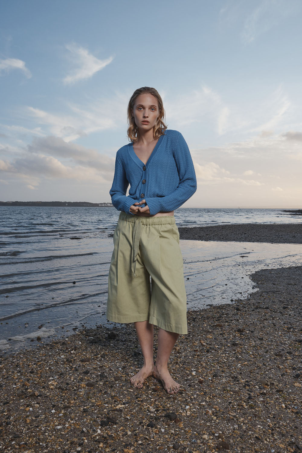 Woman wearing a blue cardigan and light green shorts standing on a beach with water and sky in the background.