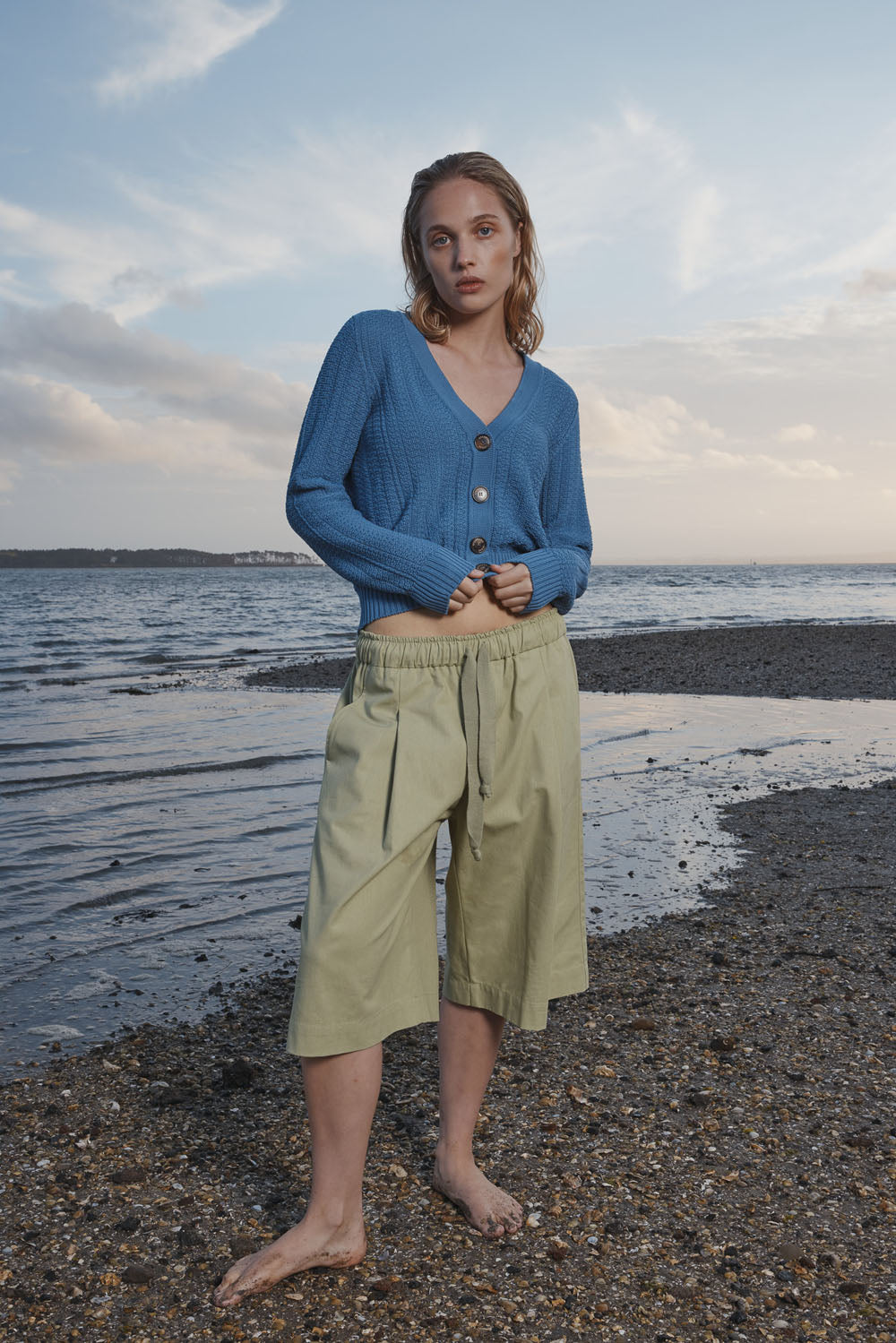 Woman wearing a blue cardigan and light green shorts standing on a beach with water and sky in the background.