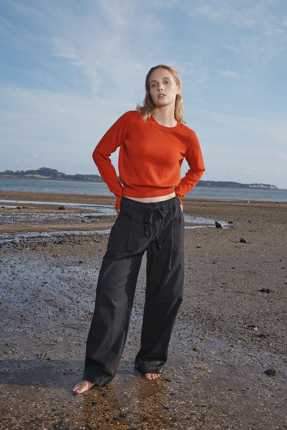 Woman wearing a red sweater and black pants standing on a beach with a blue sky.