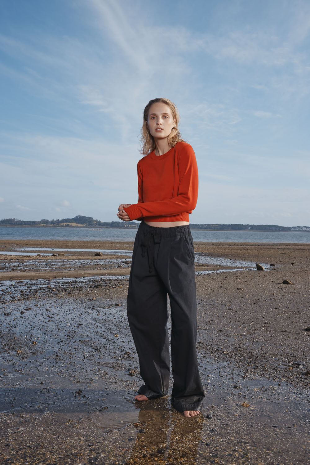 Woman in red sweater and black pants standing on a beach with a blue sky.