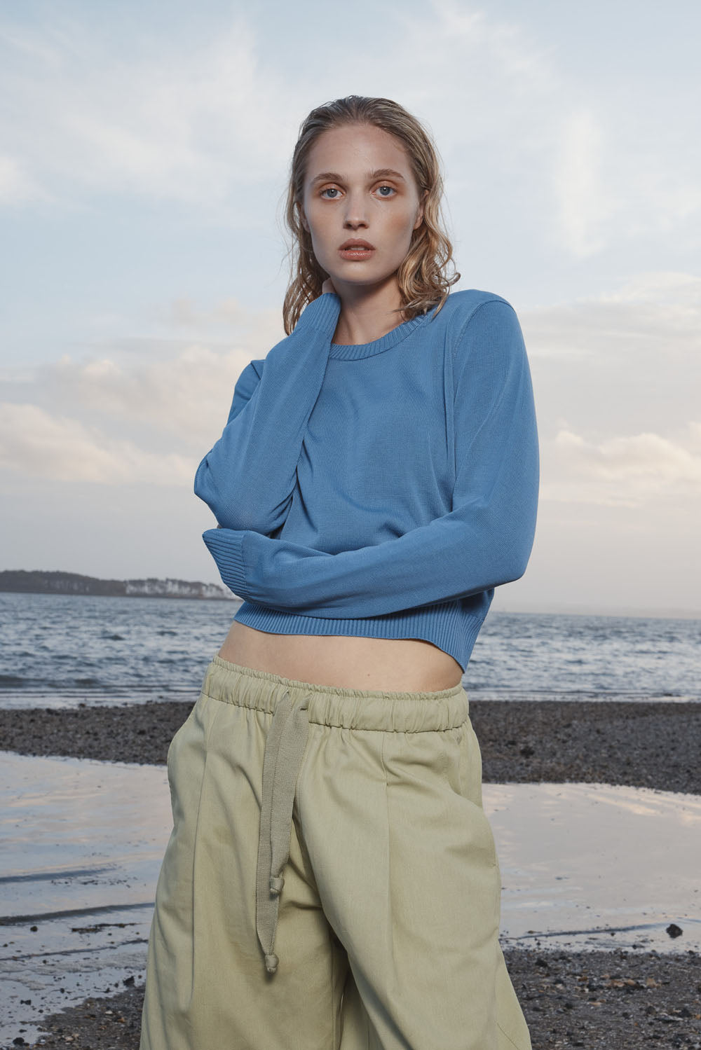 Woman wearing a blue sweater and light green pants standing on a beach with ocean and sky in the background