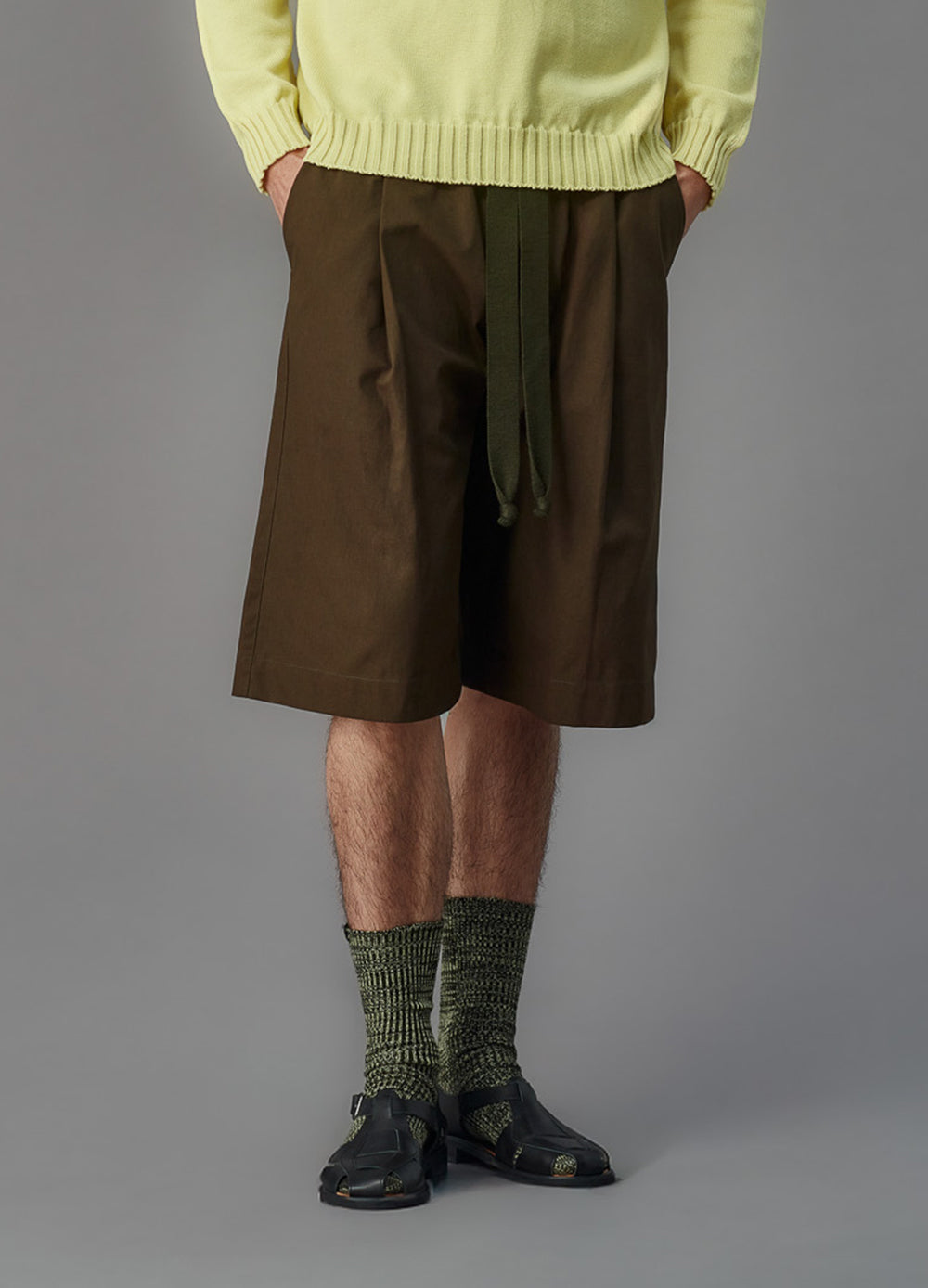 Person wearing a light green sweater, green shorts, and black shoes on a gray background