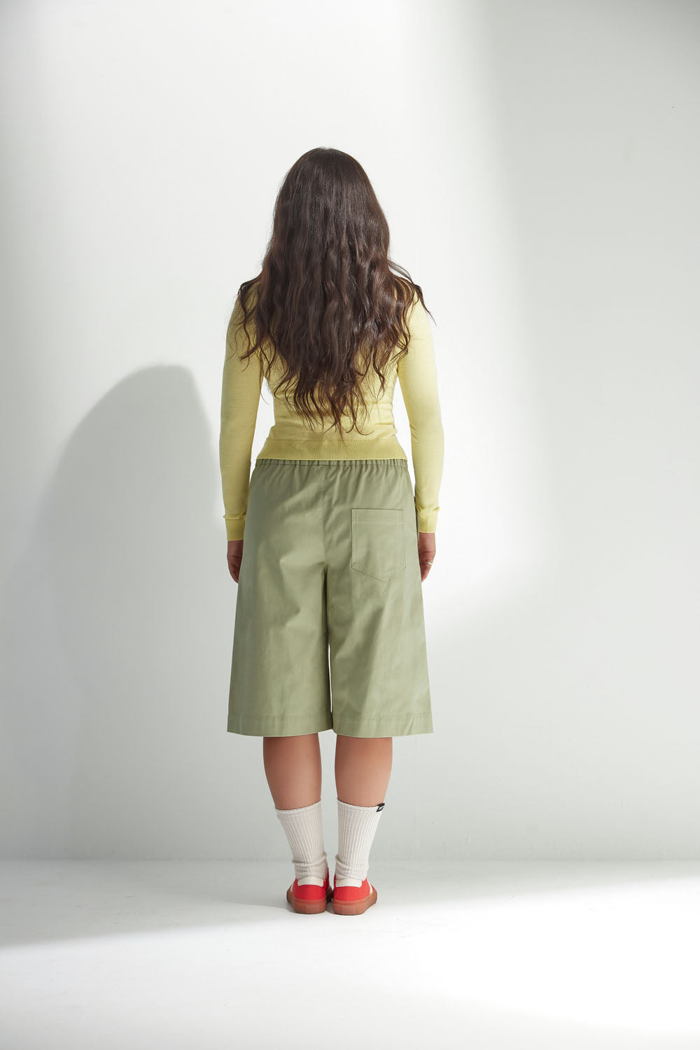 Woman wearing a yellow top and light green shorts standing against a white background with her back to the camera