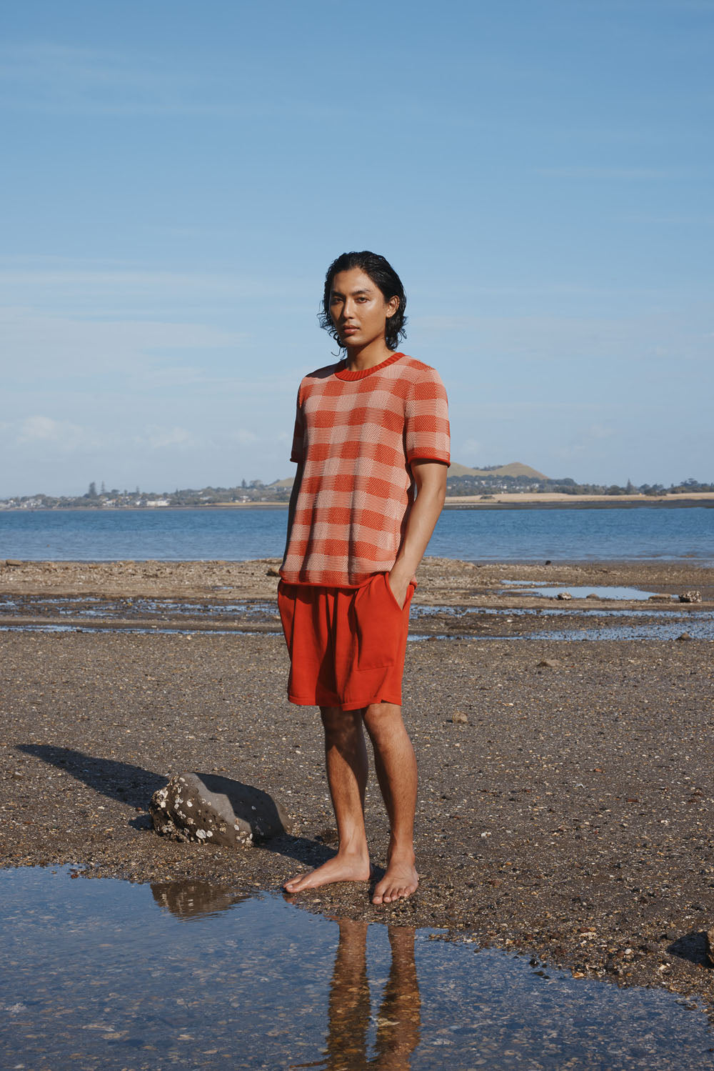 Man standing on a beach wearing a red checkered plaid shirt and red shorts.