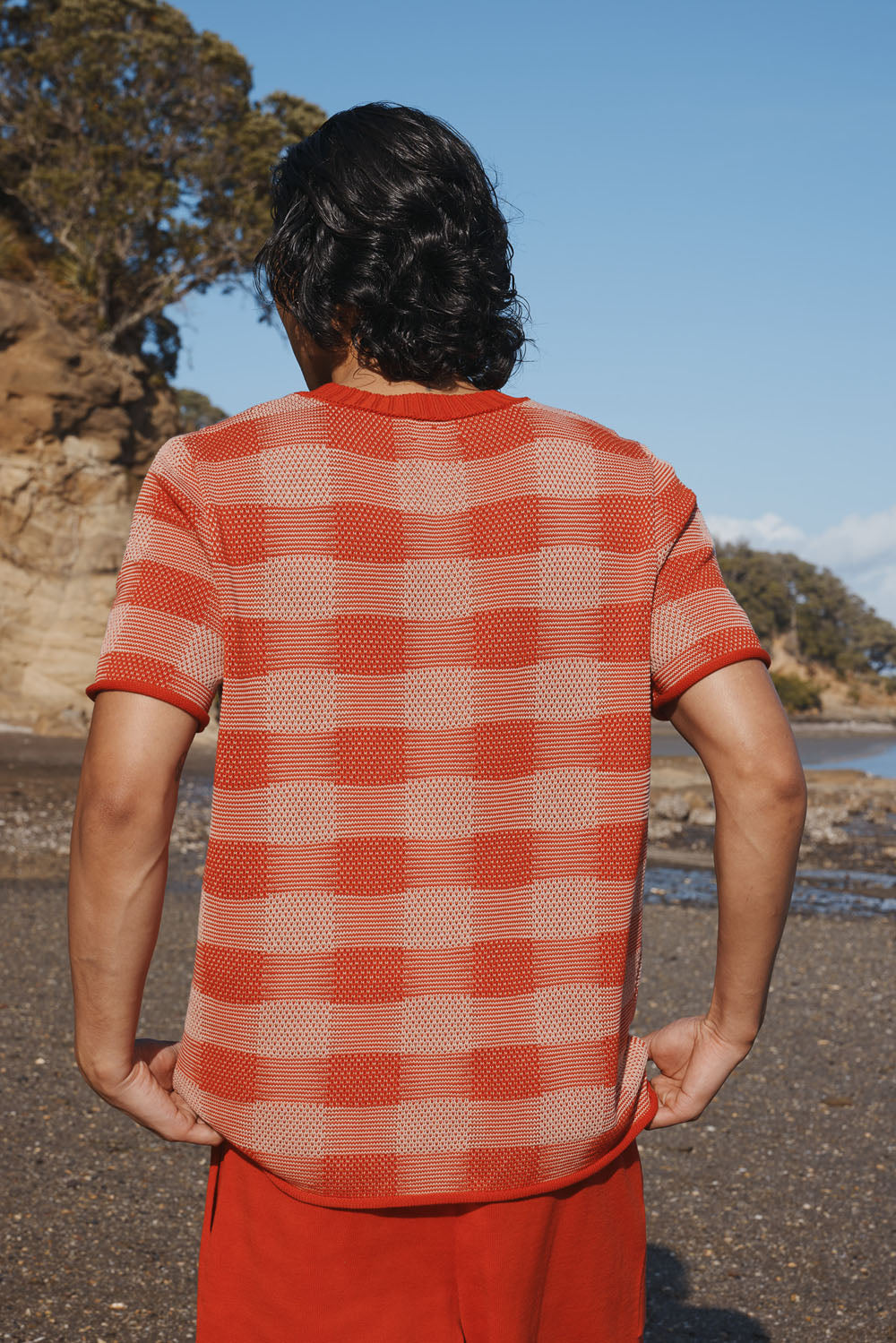 Man standing on a beach wearing a red checkered plaid shirt and red shorts with trees and sky in the background.