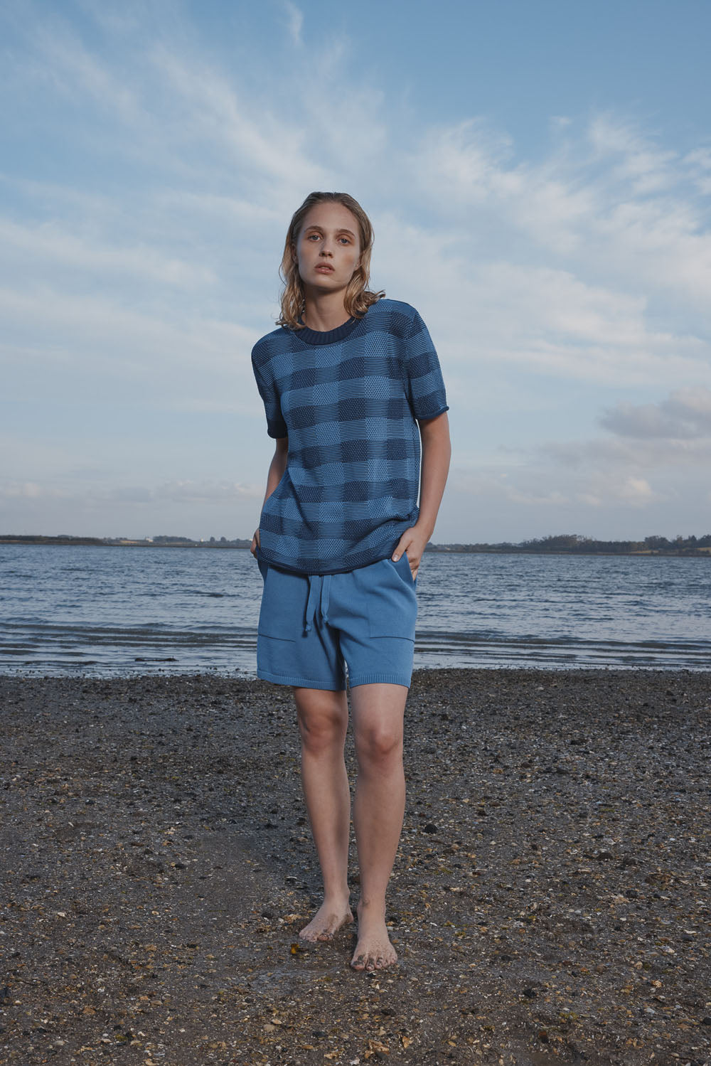 Woman wearing a blue plaid shirt and shorts standing on a beach with water and sky in the background.