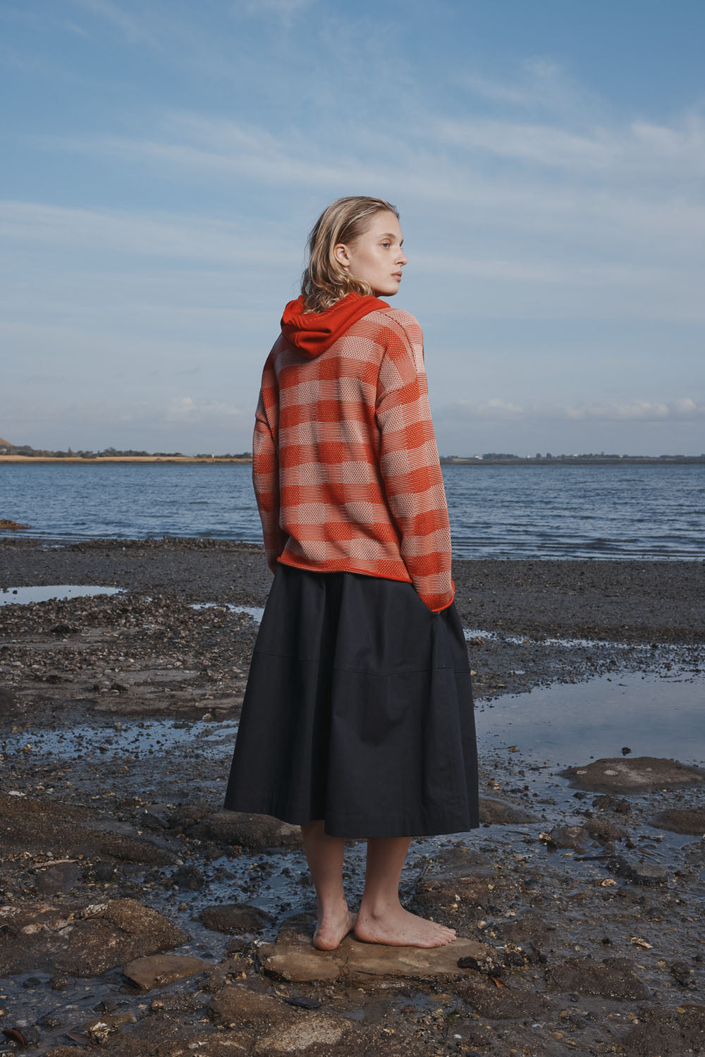 Woman wearing a red and white checkered sweater with black skirt standing on a rocky beach.