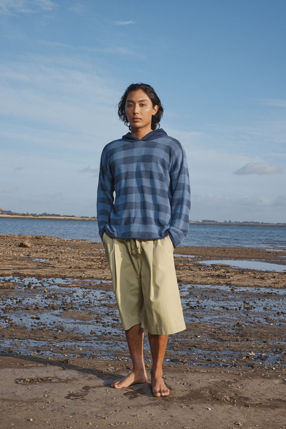 Man wearing a blue plaid sweater and light green pants standing on a beach with a clear sky.