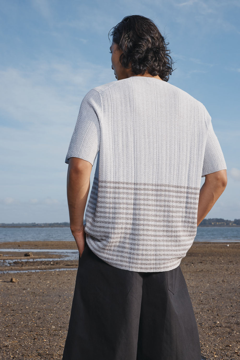 Man wearing a light gray t-shirt with a patterned back standing on a beach.