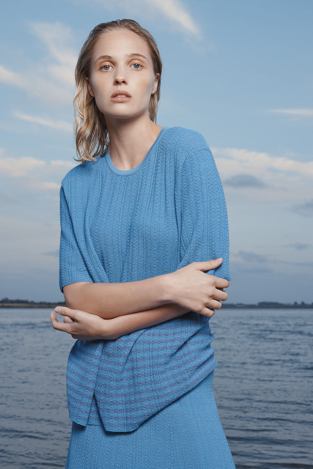 Woman in a knit blue t-shirt and skirt standing by a body of water with a cloudy blue sky.