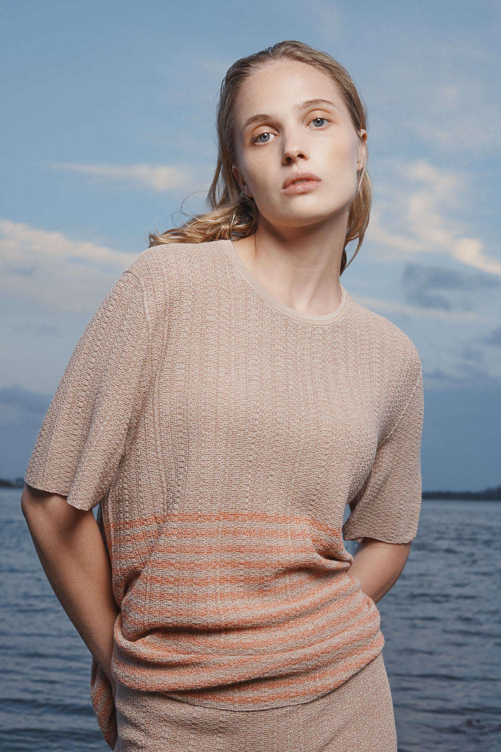 Woman wearing a taupe t-shirt standing by a body of water with a blue sky.