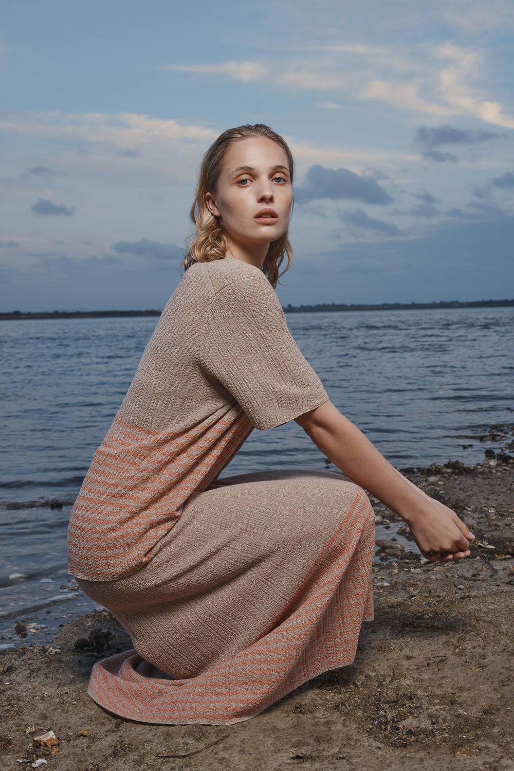 Woman wearing a taupe t-shirt crouching by a body of water with a dark blue sky.