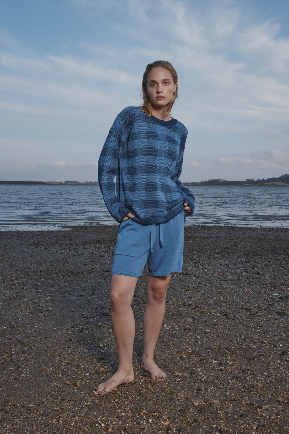 Woman wearing a blue and white checkered sweater and shorts standing on a beach with water and sky in the background.