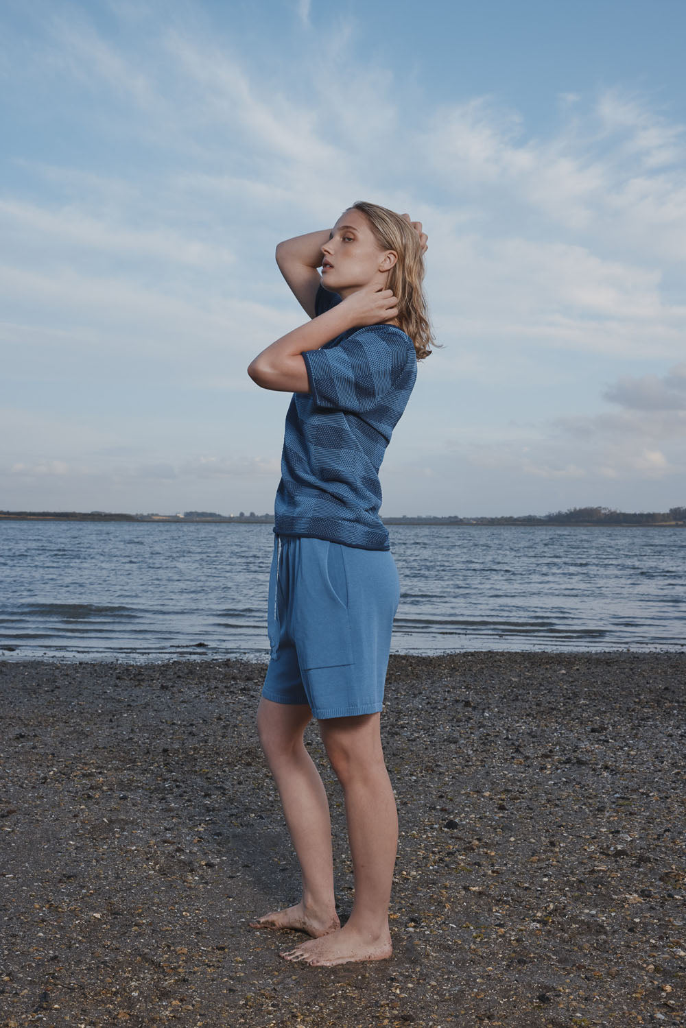Woman wearing a blue and white checkered sweater and shorts standing on a beach with water and sky in the background.