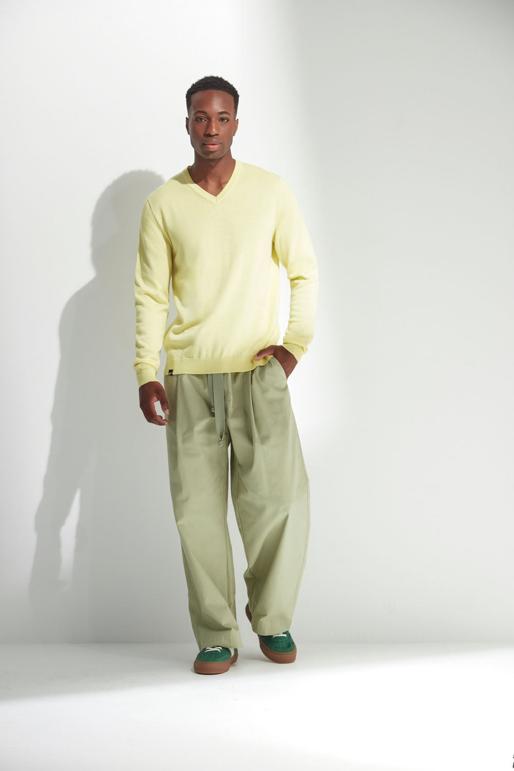 Man wearing a light yellow sweater and green pants standing against a white background