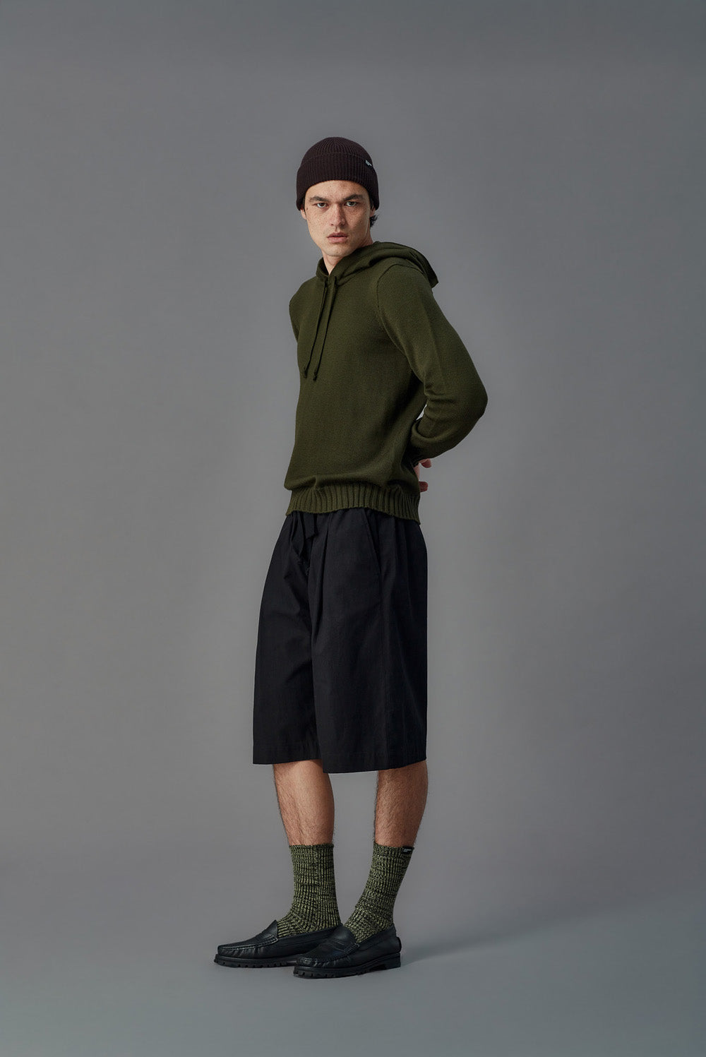 Person wearing an olive green hoodie, black shorts, and dark shoes on a gray background