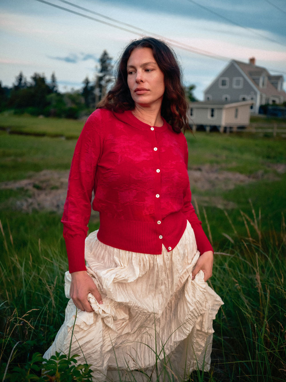 Woman wearing a red cardigan and white skirt standing in a grassy field with a house in the background.