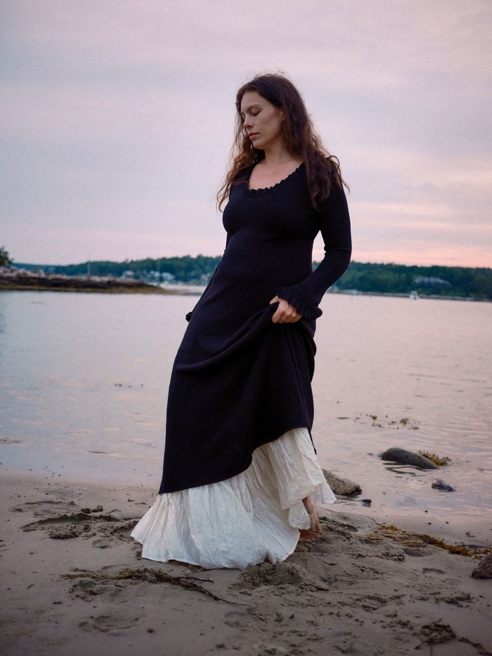 Woman in a black dress and white skirt standing on a beach at sunset.