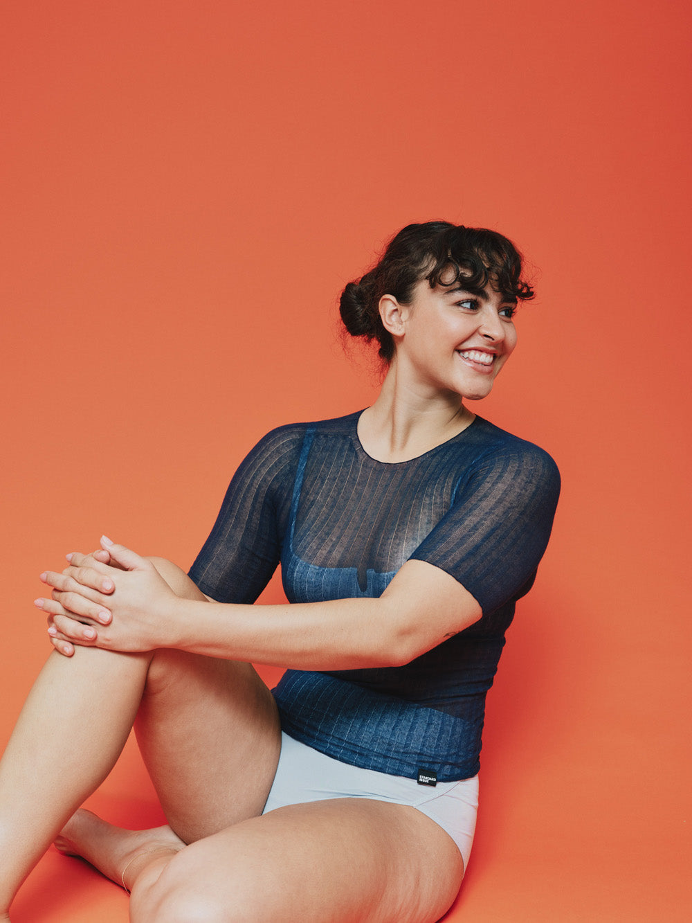 Woman sitting on an orange background wearing a blue sheer tulle top and white underwear.