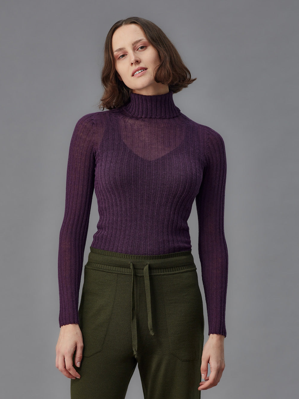 Woman wearing a purple ribbed turtleneck tulle and green pants against a gray background