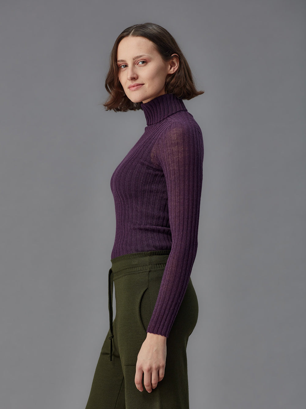 Woman wearing a purple ribbed turtleneck tulle and green pants against a gray background