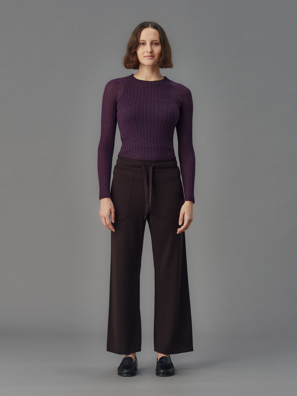 Woman wearing a purple ribbed merino tulle and dark pants on a gray background