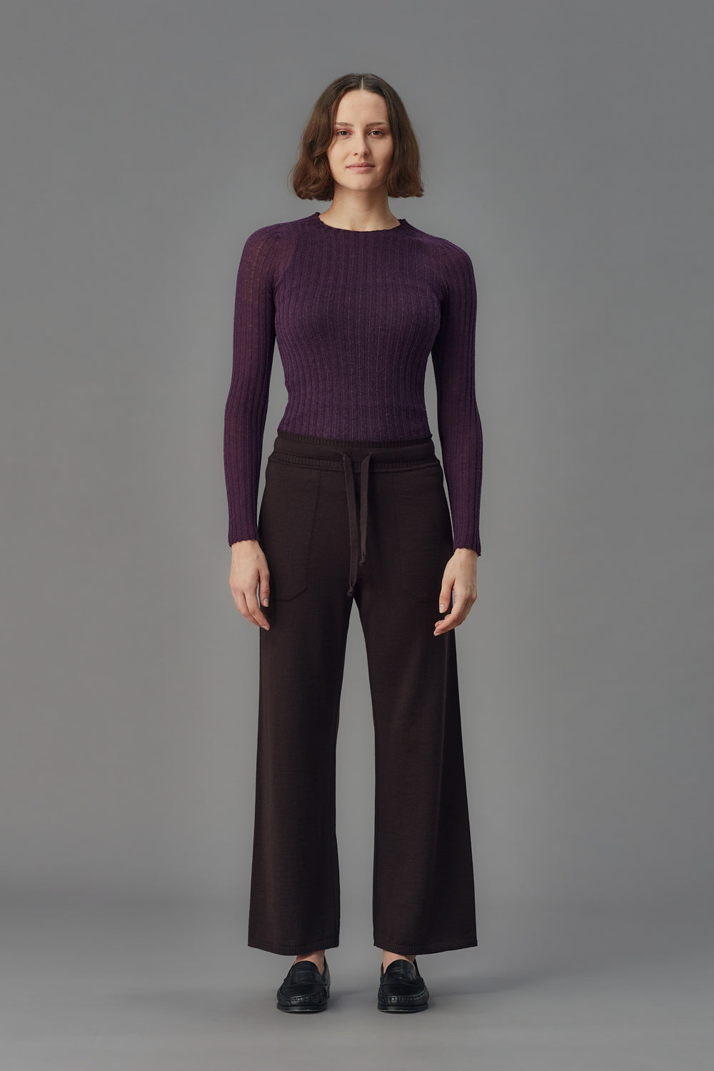 Person wearing a purple ribbed merino tulle and dark pants on a gray background