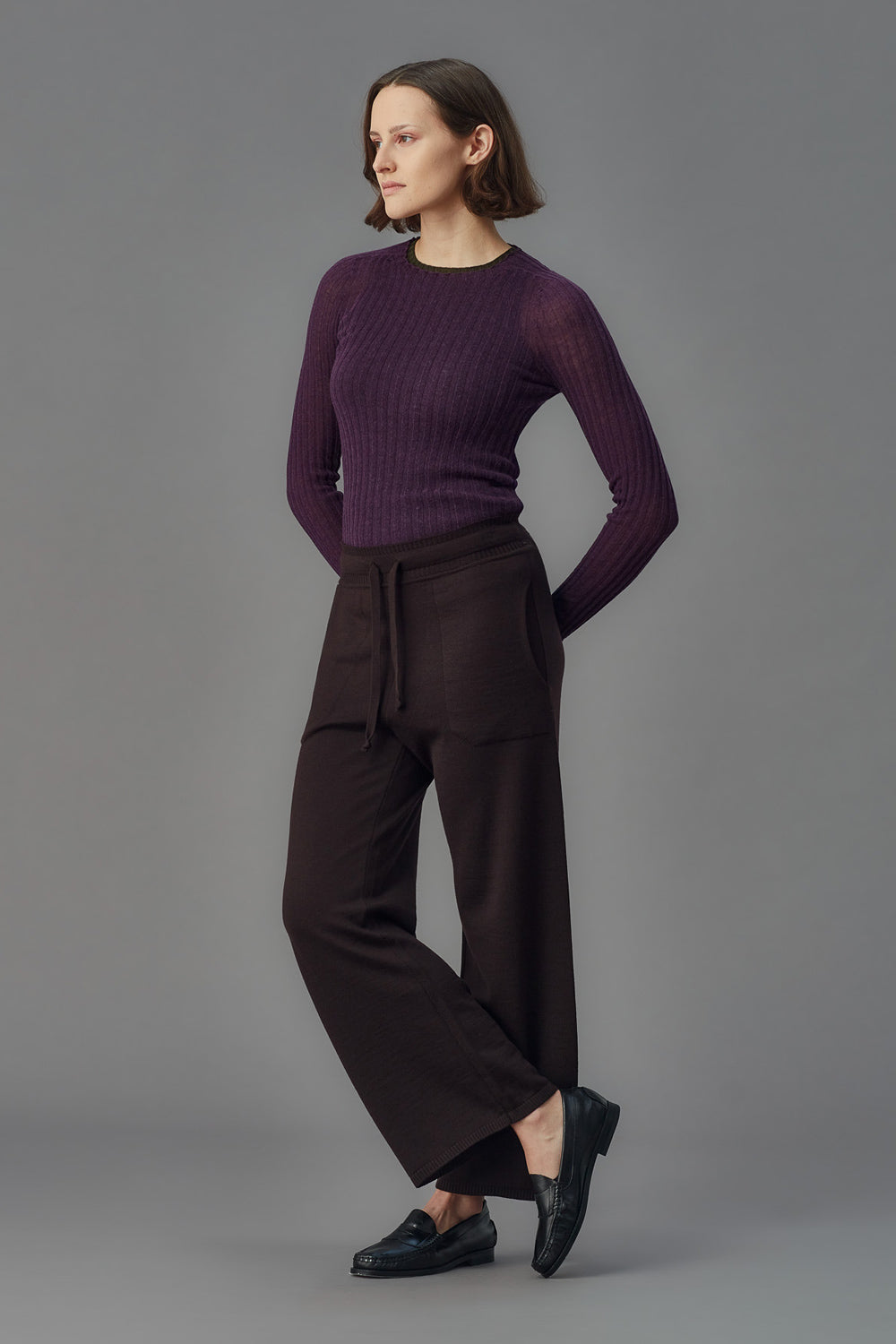 Person wearing a purple ribbed merino tulle and dark pants on a gray background