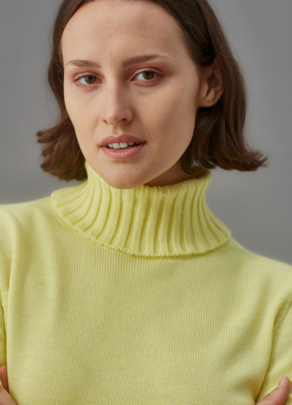 Woman wearing a light yellow funnel sweater and brown skirt against a gray background