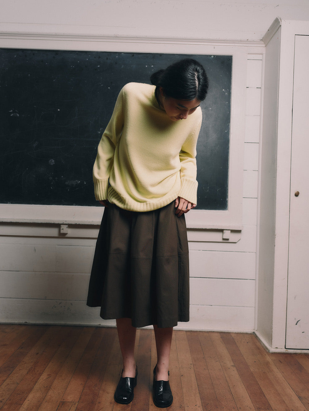 Woman in a light yellow cashmere sweater and dark skirt standing in front of a blackboard.