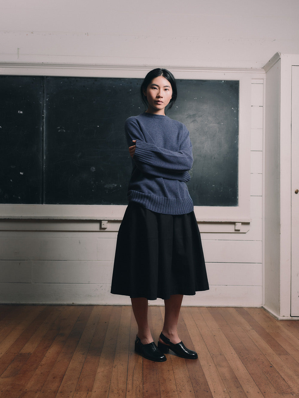 Woman wearing a blue cashmere sweater and black skirt standing in front of a chalkboard.