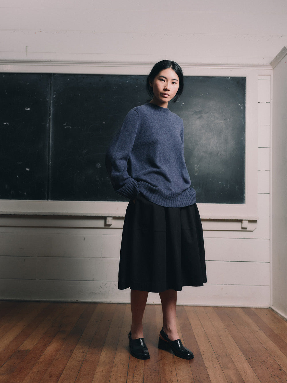 Woman wearing a blue cashmere sweater and black skirt standing in front of a chalkboard.